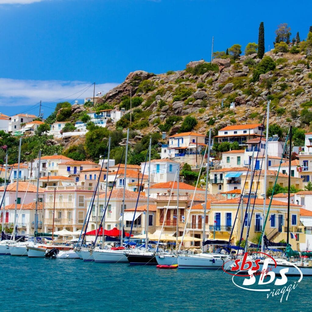 Case colorate con tetti arancioni fiancheggiano una collina dietro un porto turistico pieno di barche a vela, catturando il fascino di un'avventura del Tour Grecia. Il logo di SBS Viaggi è visibile nell'angolo in basso a destra.