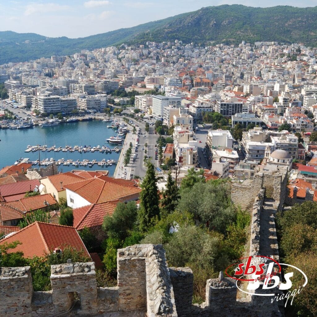Una città costiera con un porto turistico, edifici dai tetti rossi e uno storico muro di pietra in primo piano, sullo sfondo di colline e un denso sviluppo urbano: scopri Salonicco durante il tuo prossimo tour nelle Terre di Alessandro Magno.