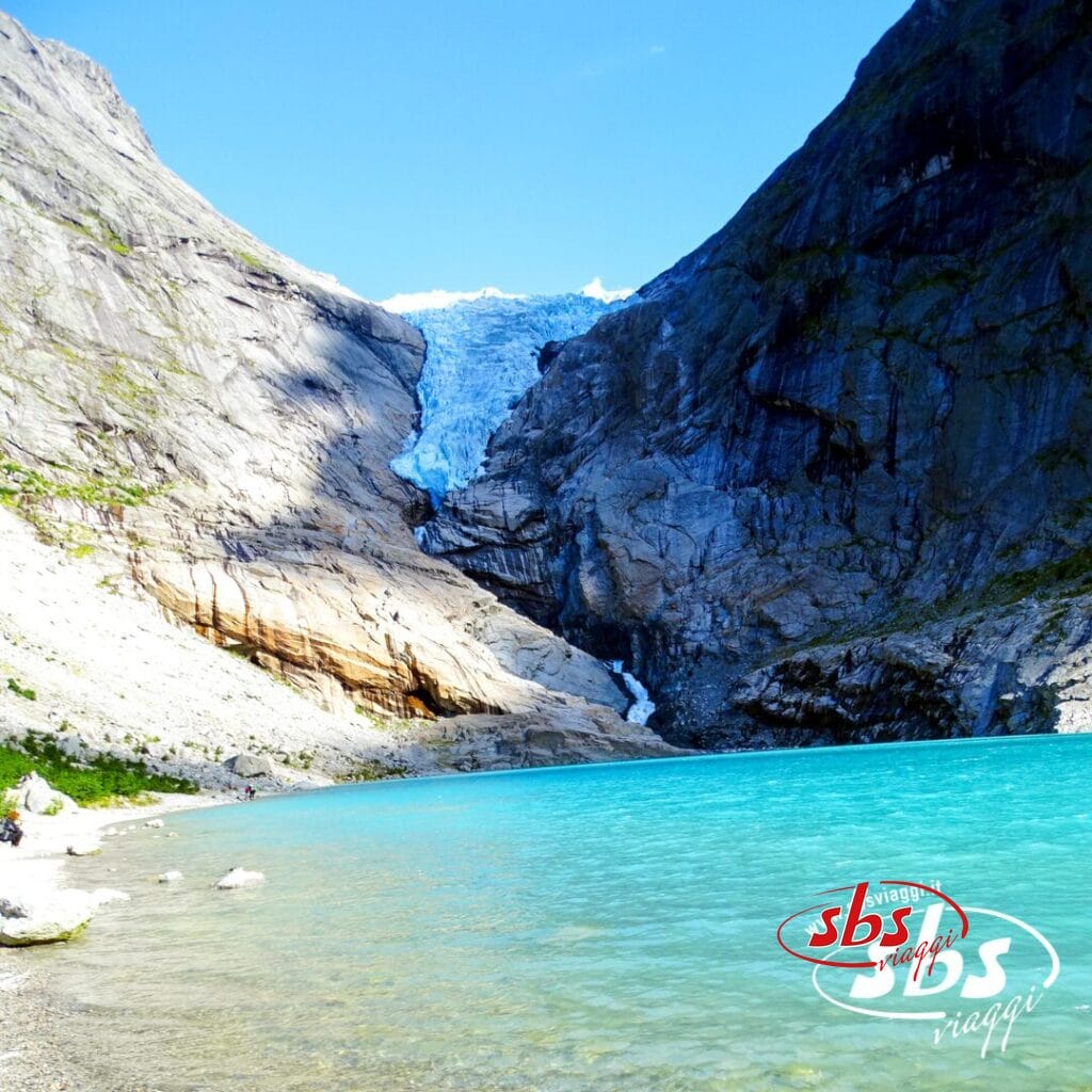 Un lago di un azzurro brillante si trova sotto un ghiacciaio incastonato tra ripide montagne rocciose e sotto un cielo azzurro e limpido, creando uno spettacolo naturale degno dei Fiordi.