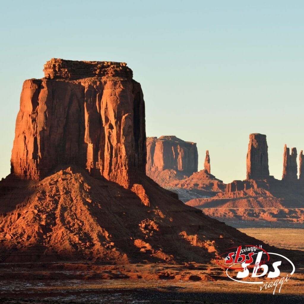 Le colline e le altopiani della Monument Valley sono illuminate dalla luce del sole, proiettando lunghe ombre sul paesaggio desertico; nell'angolo in basso a destra appare il logo della compagnia di viaggi Explorer 1924.