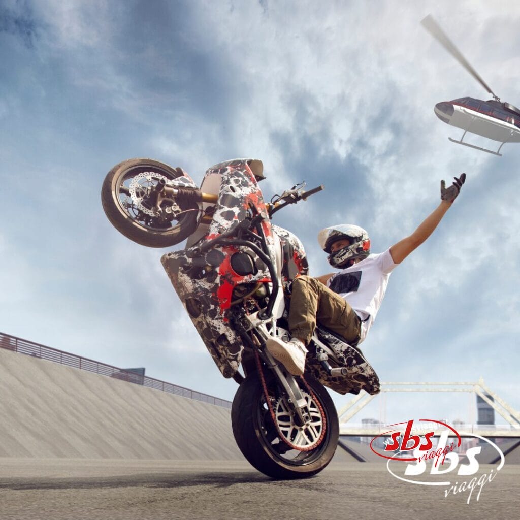 Un motociclista esegue un'audace impennata su una rampa con una mano alzata, mentre un elicottero vola sullo sfondo. L'immagine, con il logo 