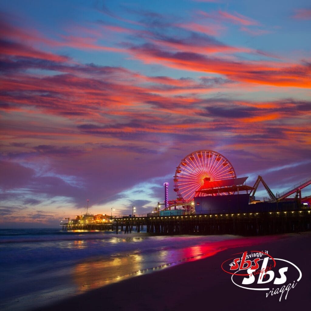 Una ruota panoramica su un molo di Los Angeles è illuminata al tramonto, con nuvole rosa e arancioni vivaci nel cielo e l'oceano in primo piano. Il logo di SBS Viaggi, ispirato a West 1924, appare nell'angolo.