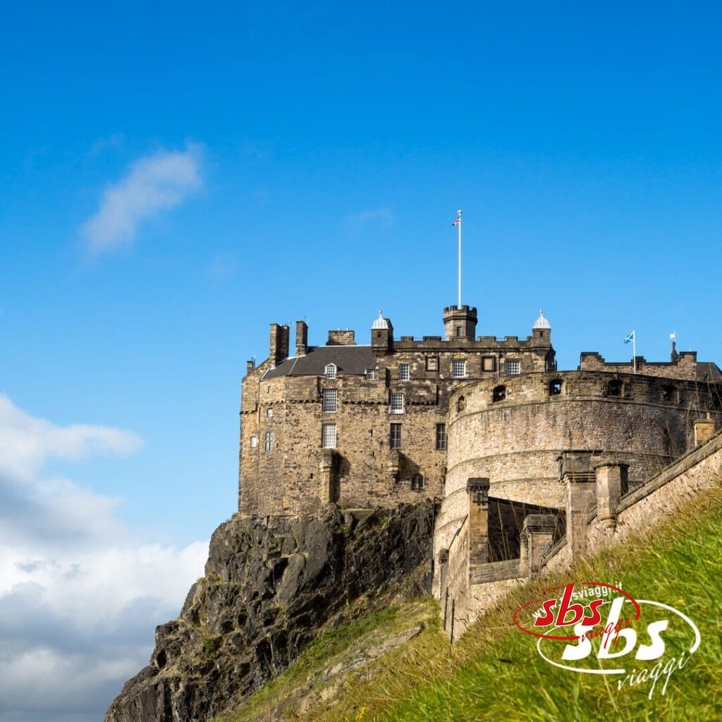Un castello di pietra si erge sulla cima di una collina rocciosa sotto un cielo azzurro e limpido sull'isola di Scozia, con una bandiera che sventola dalla sua torre e un pendio erboso in primo piano, incarnando il fascino senza tempo dei castelli scozzesi.