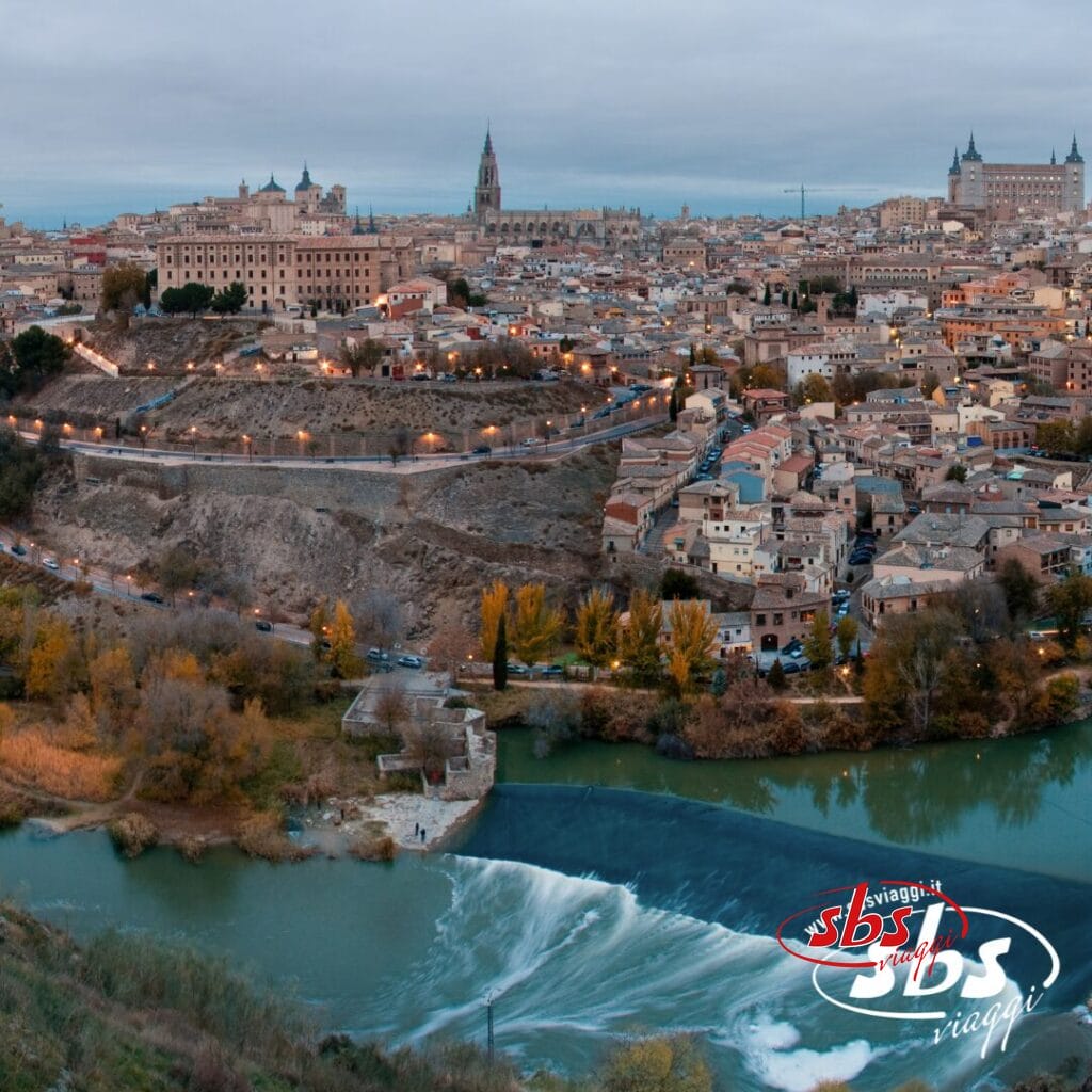 Vista aerea di una città storica che riflette il fascino della Spagna Medievale, con un fiume che scorre in primo piano. Lo skyline, ideale per un tour avvincente, presenta antichi edifici e un'imponente cattedrale sotto un cielo coperto.