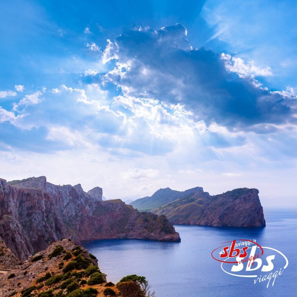 Costa rocciosa con ripide scogliere e mare azzurro sotto un cielo parzialmente nuvoloso, con raggi di sole che filtrano: uno scenario ideale per un tour a Capo Nord. Il logo