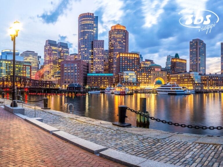Lo skyline della città al tramonto riflesso nell'acqua, con moderni grattacieli, una barca attraccata e una passerella in mattoni in primo piano: perfetto per la tua esperienza di viaggio sulla costa orientale di Metropoli.