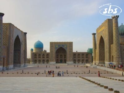 Una grande piazza aperta con gruppi di persone, circondata da tre grandi edifici islamici riccamente decorati con cupole blu e ingressi ad arco: un luogo iconico lungo la storica Via Della Seta consigliata da Francorosso.