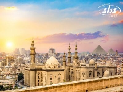 Una vista panoramica del Cairo con le moschee storiche in primo piano, le Piramidi di Giza visibili in lontananza e il maestoso Nilo che serpeggia sotto un cielo colorato al tramonto.