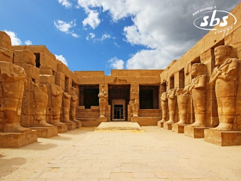 Cortile di un antico tempio egizio al Cairo, con grandi statue in pietra che costeggiano il vialetto d'ingresso, mura di arenaria e un cielo parzialmente nuvoloso in alto, vicino al maestoso Nilo.