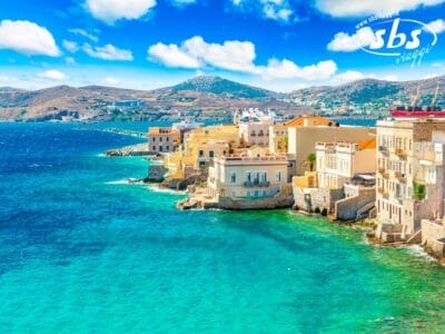 Colorati edifici sul mare costeggiano la costa turchese di una città mediterranea, con le montagne sullo sfondo e un cielo azzurro. Perfetto per un tour in Grecia o una rilassante fuga in Grecia mare. Il logo "sbs viaggi" appare nell'angolo in alto a destra.