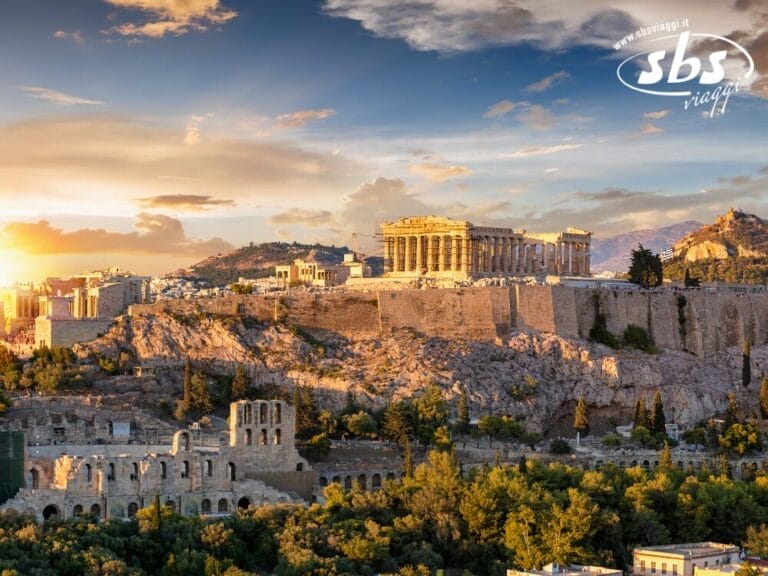 L'Acropoli di Atene con il Partenone illuminato dal tramonto, circondato da antiche rovine e vegetazione, sotto un cielo parzialmente nuvoloso: una tappa obbligata di qualsiasi tour della Grecia o viaggio attraverso la Grecia Classica.