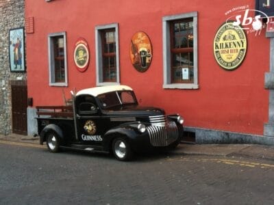 Un camioncino nero d'epoca della Guinness è parcheggiato su una strada acciottolata, fuori da un pub, con insegne di birra e una ricetta di cocktail irlandese dipinta sulla parete esterna rossa.