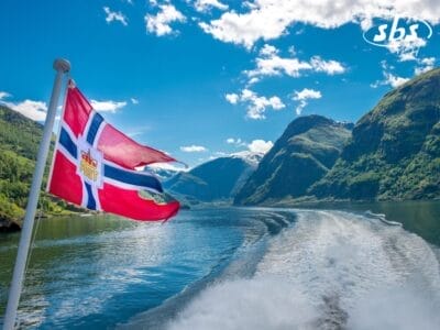 Una bandiera norvegese sventola sul retro di una barca che naviga nella Terra dei Fiordi, con montagne mozzafiato e cieli azzurri che dipingono uno sfondo perfetto per la Norvegia.
