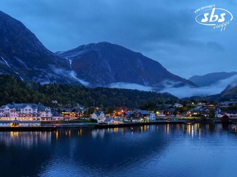 Graziosa cittadina in Norvegia, con edifici colorati in riva a un lago calmo, circondata da montagne e sotto un cielo azzurro nuvoloso al tramonto: qui la Magia dei Fiordi prende vita.