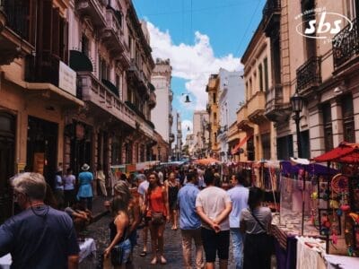 La gente passeggia lungo un vivace mercato all'aperto di Buenos Aires, in Argentina, fiancheggiato da bancarelle colorate e vecchi edifici sotto un cielo azzurro e nuvole.