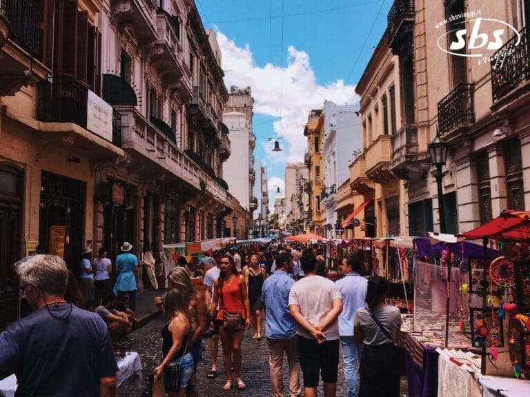 La gente passeggia lungo un vivace mercato all'aperto di Buenos Aires, in Argentina, fiancheggiato da bancarelle colorate e vecchi edifici sotto un cielo azzurro e nuvole.
