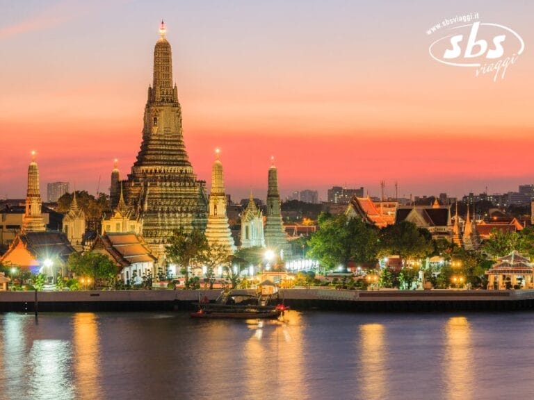 Complesso di templi illuminato al tramonto lungo un fiume, con un'alta pagoda come punto focale e gli edifici della città visibili sullo sfondo: uno scenario iconico della Thailandia, perfetto per il tuo prossimo Grantour o tour in Thailandia.