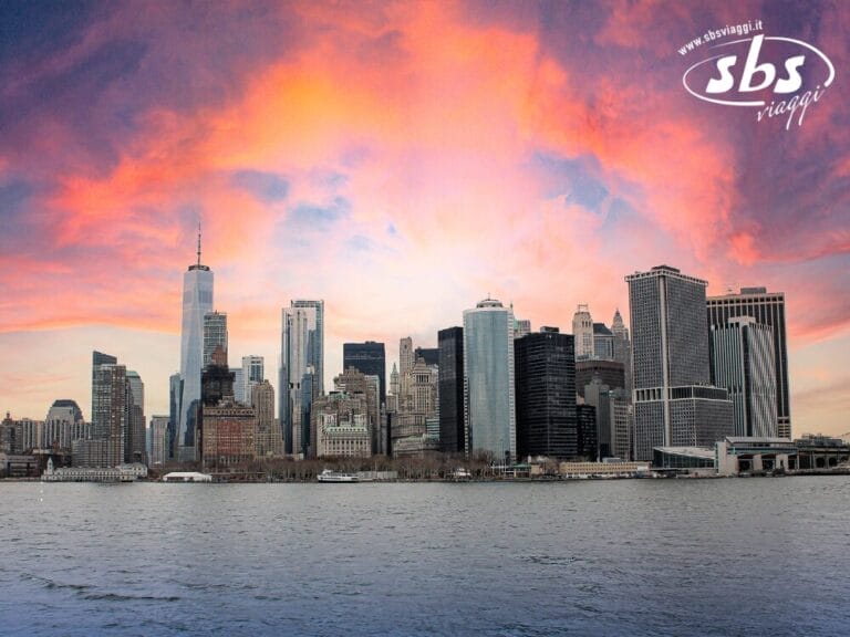 Lo skyline di New York visto dall'acqua al tramonto, con i suoi moderni grattacieli e un cielo colorato, perfetto per visitare la città. Il logo "sbs viaggi" è visibile nell'angolo in alto a destra.