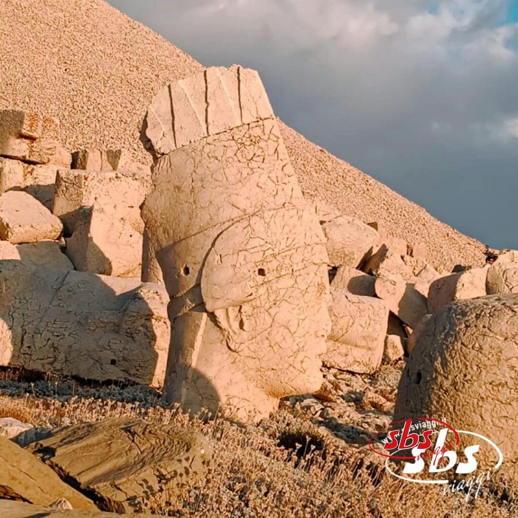 Una grande e antica statua in pietra raffigurante una testa adagiata su un terreno roccioso sul monte Nemrut, con frammenti di pietra sparsi e uno sfondo in pendenza sotto un cielo parzialmente nuvoloso: uno spettacolo iconico da ammirare durante un Viaggio in Turchia.