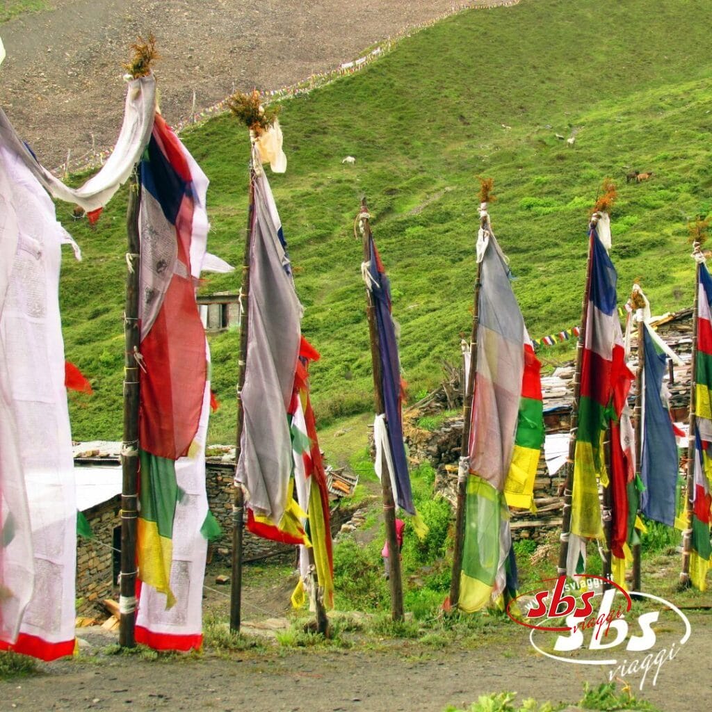 Bandiere di preghiera colorate su pali alti sono disposte in fila su un pendio erboso vicino alle strutture, con la Valle degli Dèi e le maestose montagne sullo sfondo: uno scenario perfetto per chi desidera visitare il Nepal.