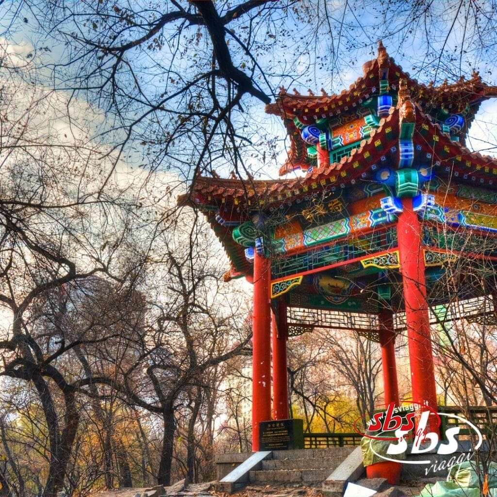 Un padiglione cinese tradizionale con colonne rosse ornate e un tetto colorato si erge tra gli alberi spogli di un parco in autunno, catturando il fascino della Cina Classica: un luogo ideale per il tuo prossimo tour in Cina.