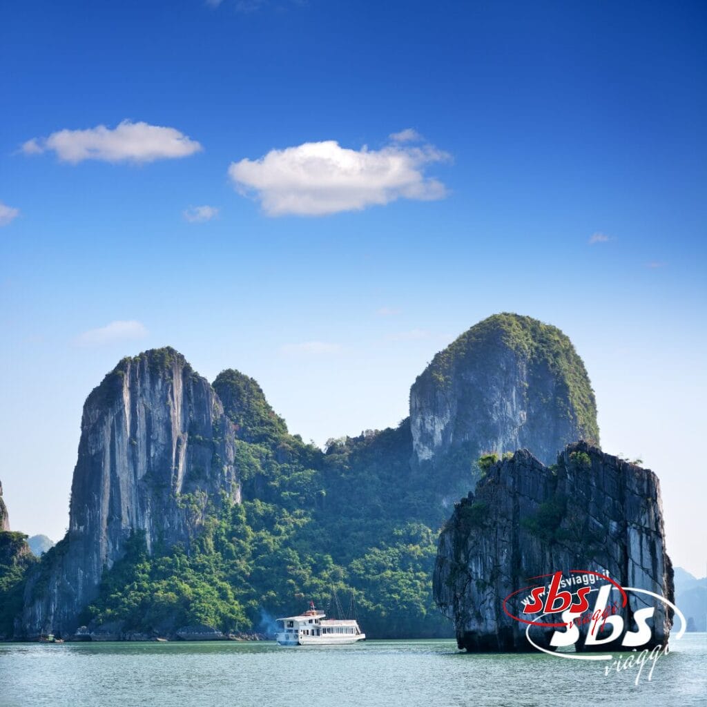Una barca bianca naviga tra isole calcaree verdi e formazioni rocciose su acque calme e azzurre sotto un cielo limpido: uno scenario indimenticabile di un tour in Vietnam. Il logo