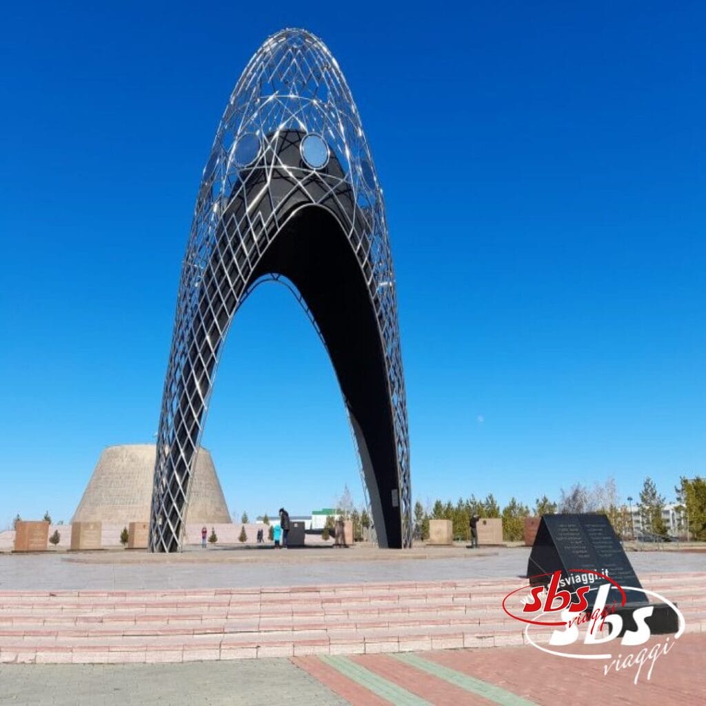 Un grande monumento metallico ad arco si erge su una piazza pavimentata sotto un cielo azzurro e limpido, con alcune persone nelle vicinanze e una targa informativa in primo piano: una tappa obbligata per qualsiasi Tour Kazakistan o viaggio Kazakistan.