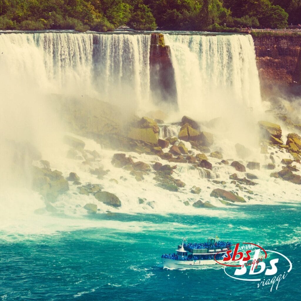 Un battello turistico pieno di persone si avvicina alla base della grande cascata del Niagara, circondato da nebbia, alberi e terreno roccioso. Il logo 