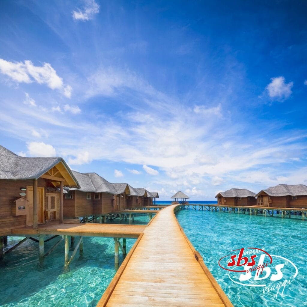 Passerella in legno sulle acque cristalline che conduce ai bungalow sull'acqua alle Maldive, sotto un cielo luminoso con nuvole sparse; logo