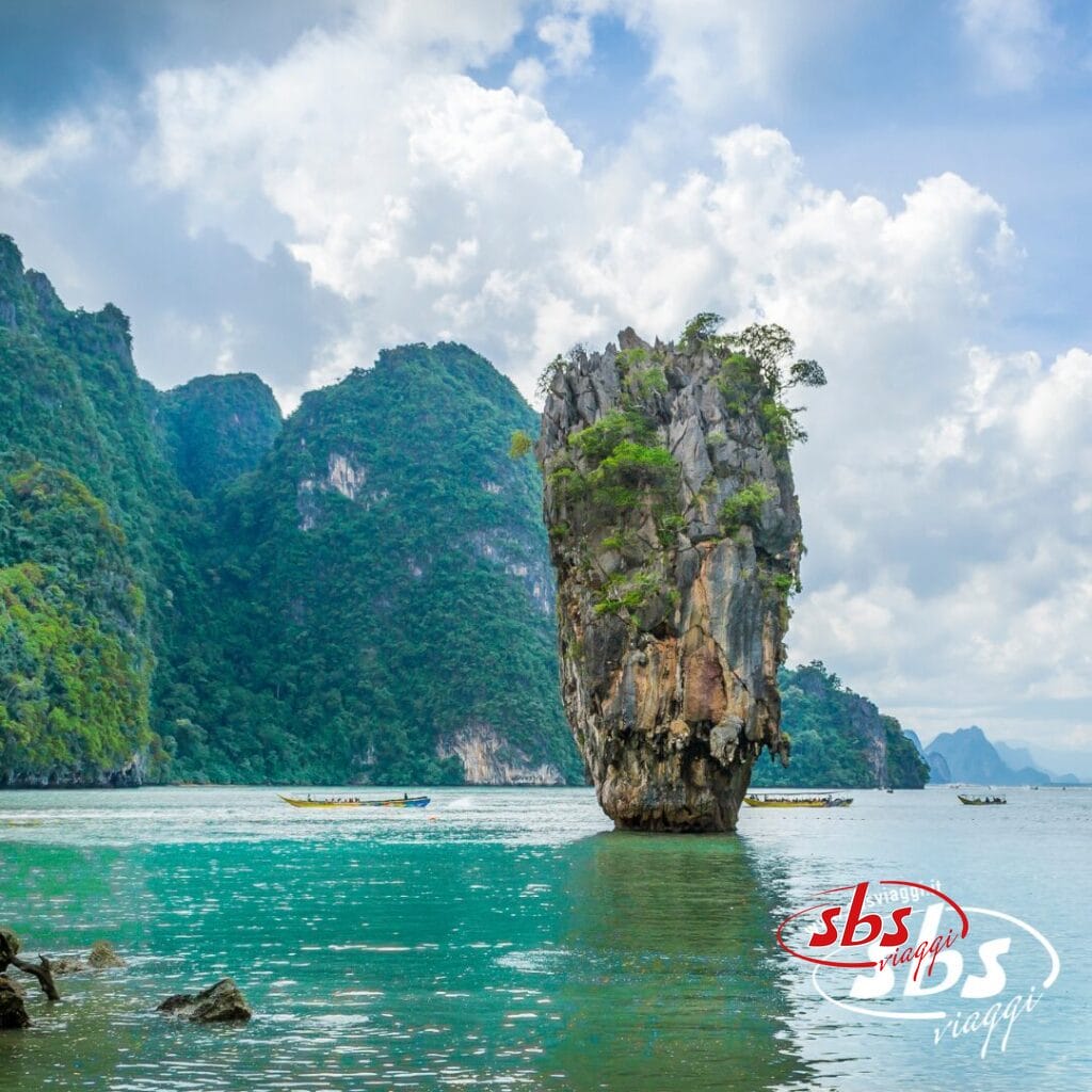 Un'alta isola rocciosa ricoperta di verde si erge dalle acque turchesi, con le montagne sullo sfondo, catturando la magia della Thailandia Magica e la serena bellezza vicino a Phuket.