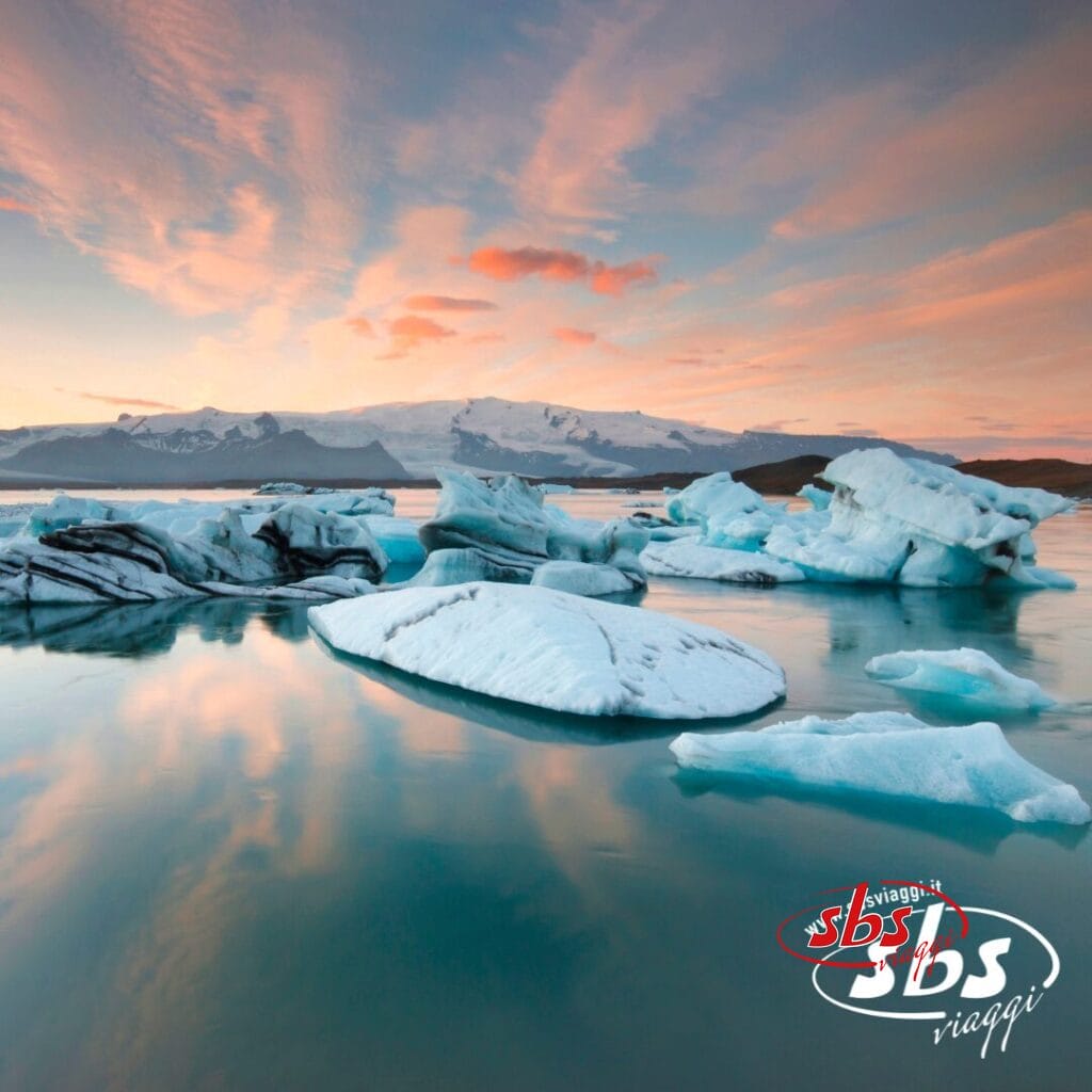 Iceberg che galleggiano in acque calme al tramonto, con montagne e un cielo colorato sullo sfondo. Con il logo 