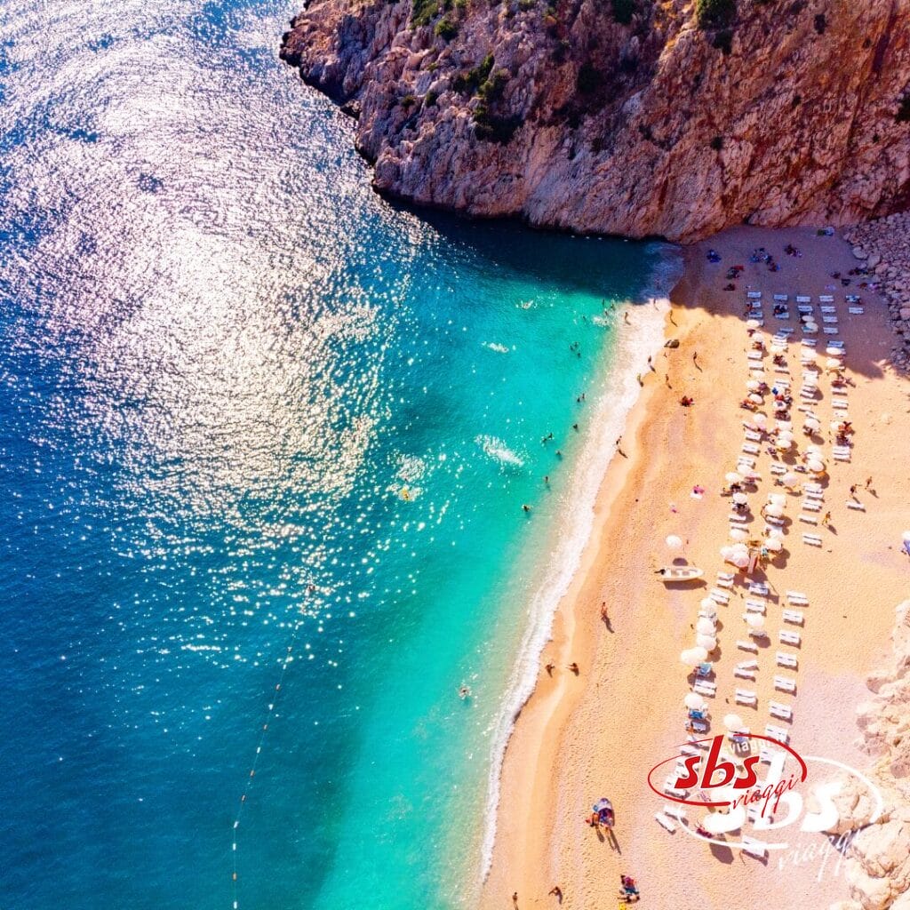 Vista aerea di una spiaggia sabbiosa con acqua turchese, lettini e ombrelloni allineati lungo la riva vicino a scogliere rocciose: uno scenario invitante da ammirare durante un tour Sapori del Mediterraneo o mentre si esplora la splendida Lykia.