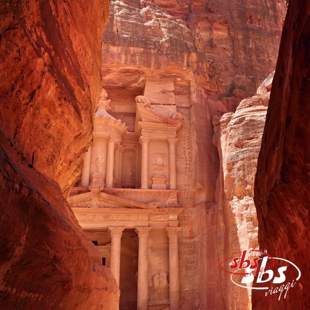 Antica facciata in pietra del Tesoro di Petra in Giordania, incorniciata da scogliere di roccia rossa, con il logo SBS Viaggi: il tuo viaggio completo per un Gran Tour indimenticabile.