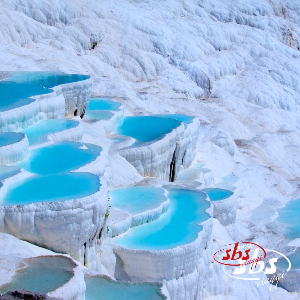 Piscine termali turchesi a gradini formate da terrazze minerali bianche a Pamukkale, in Turchia, con il logo SBS Viaggi nell'angolo in basso a destra: una tappa iconica dell'itinerario del Tour Kervansaray.