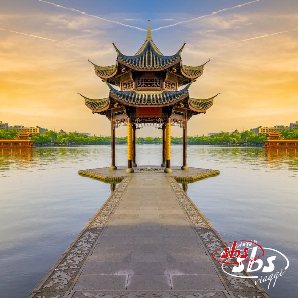 Un tradizionale padiglione cinese con un tetto decorato si erge alla fine di una passerella in pietra sopra acque calme, evocando la serena bellezza della Cina Classica. Perfetto per la tua prossima avventura con il Tour Balla coi Panda, sotto un cielo limpido e con la città sullo sfondo.