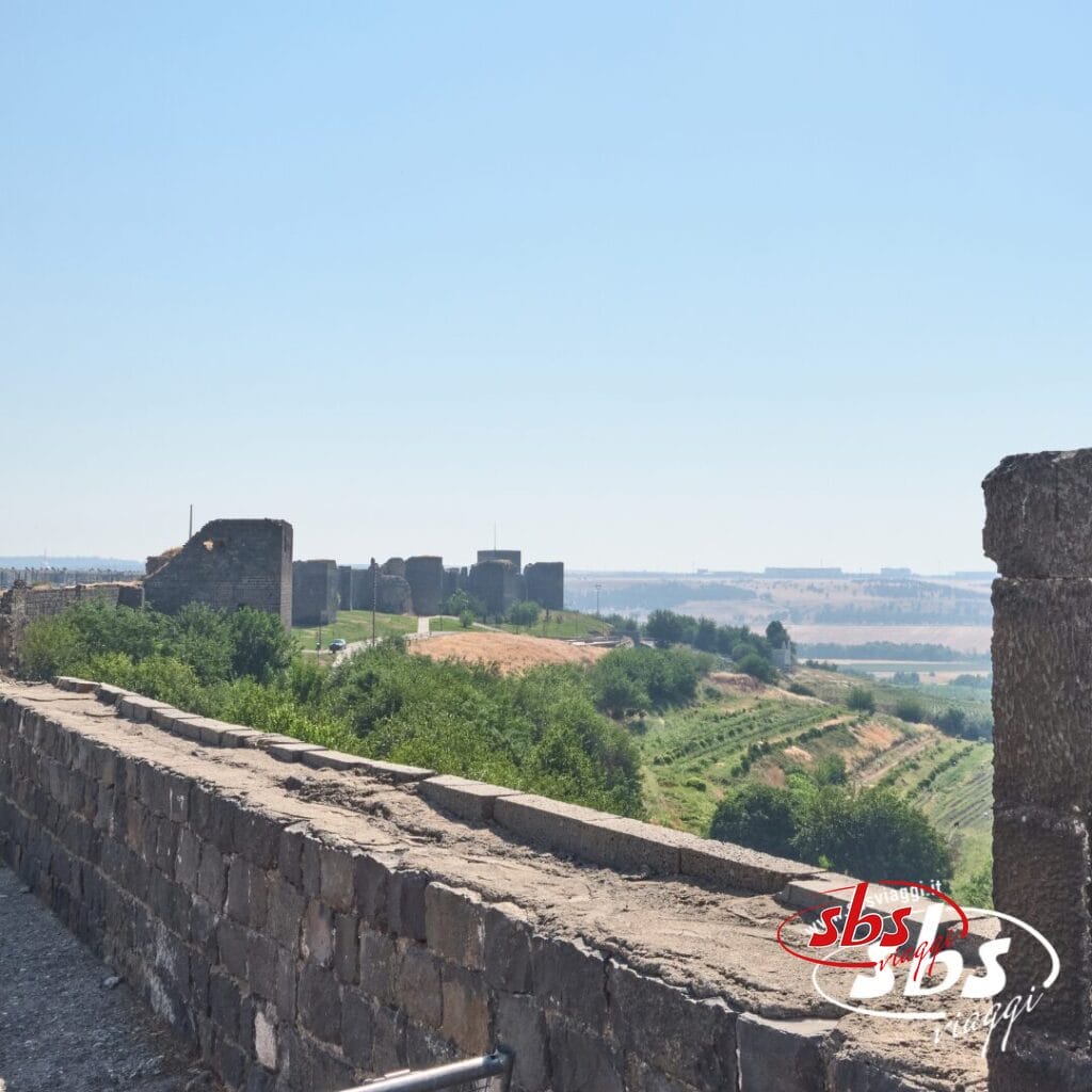 Le antiche mura di pietra della fortezza si affacciano su campi verdi e colline lontane sotto un cielo azzurro e limpido, catturando i Colori della Turchia nel tuo Viaggio Turchia.