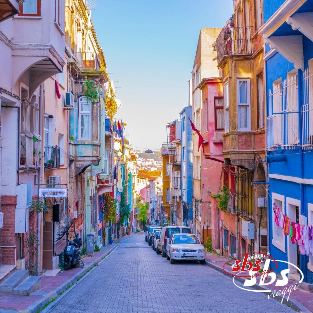 Una strada stretta e in pendenza in Cappadocia, fiancheggiata da edifici colorati e auto parcheggiate sotto un cielo limpido, con i panni stesi sulla destra. Il logo