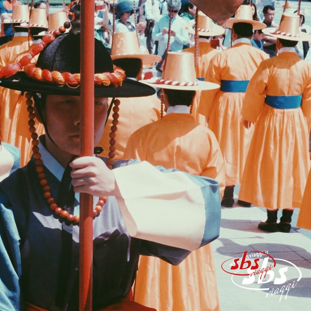 Un gruppo di persone in abiti tradizionali coreani partecipa a una sfilata durante il Tour Corea del Sud; una persona in testa tiene un palo e indossa una collana di perline e un cappello a tesa larga.