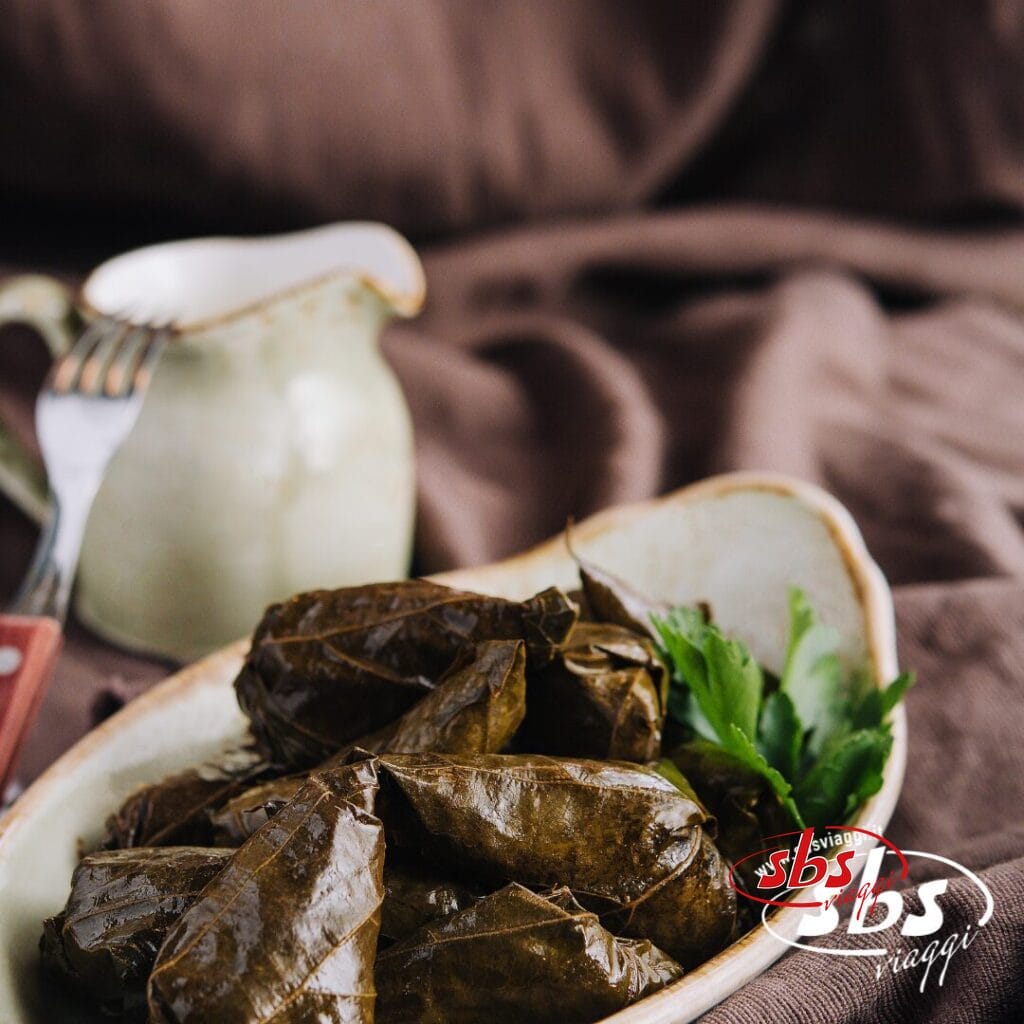 Un piatto di ceramica con foglie di vite ripiene, un piatto popolare in Azerbaigian e Georgia, guarnito con prezzemolo, con una piccola brocca e una forchetta sullo sfondo.