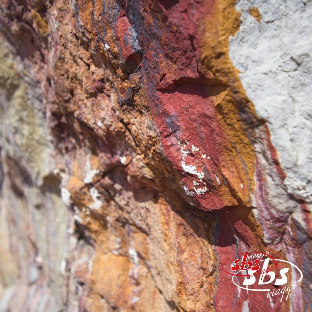 Primo piano della superficie di una roccia caratterizzata da strati di minerali rossi, gialli e bianchi nella Valle degli Dei in Nepal, con il logo 