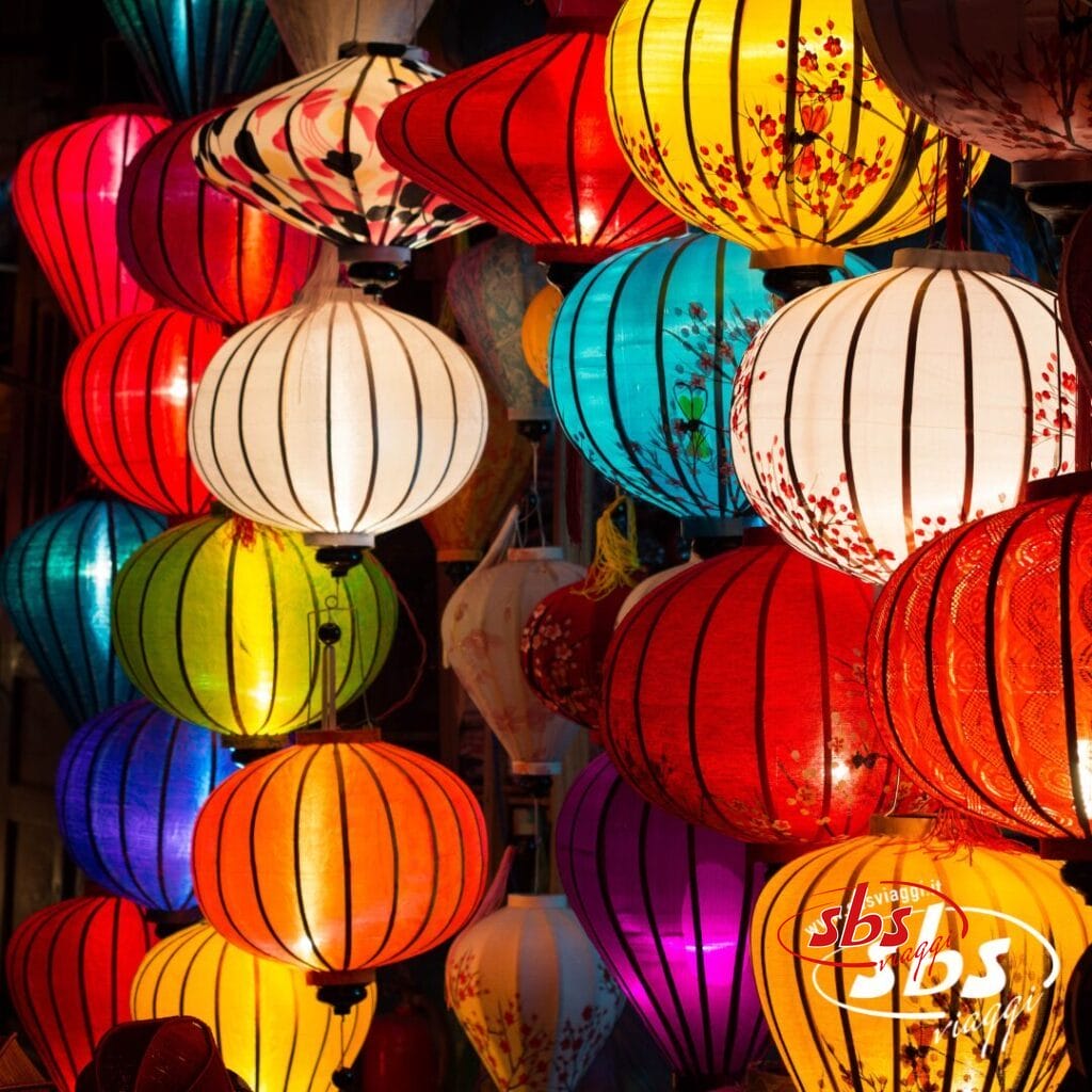 Lanterne colorate di varie forme e fantasie sono appese vicine, illuminate e creano un'atmosfera vivace: una scena iconica che si ritrova spesso durante il Gran Tour del Vietnam. L'immagine presenta la filigrana