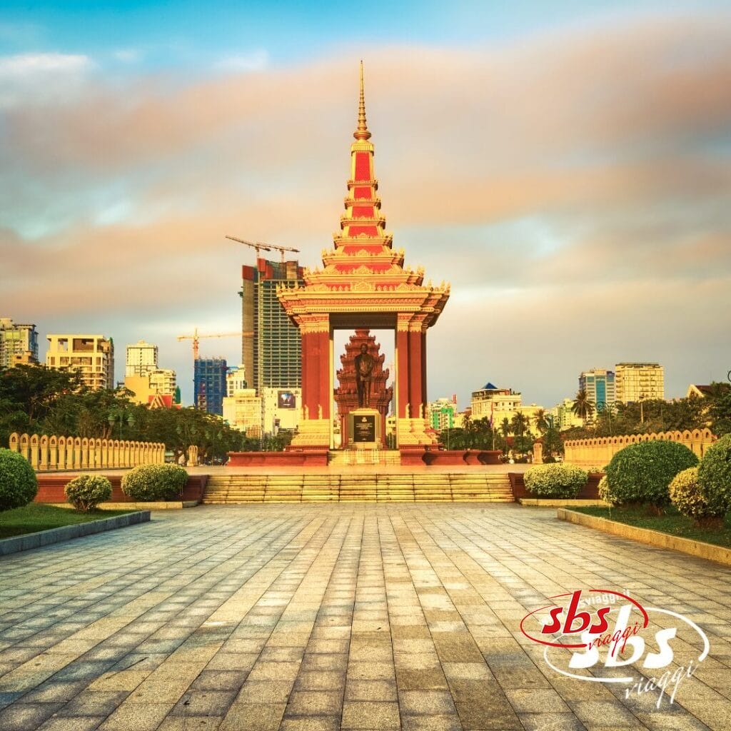 Un monumento con il tetto rosso e oro si erge in una piazza, circondato dal verde, con edifici moderni e gru sullo sfondo: un luogo suggestivo da visitare durante la tua avventura con Grantour o Tour Cambogia.