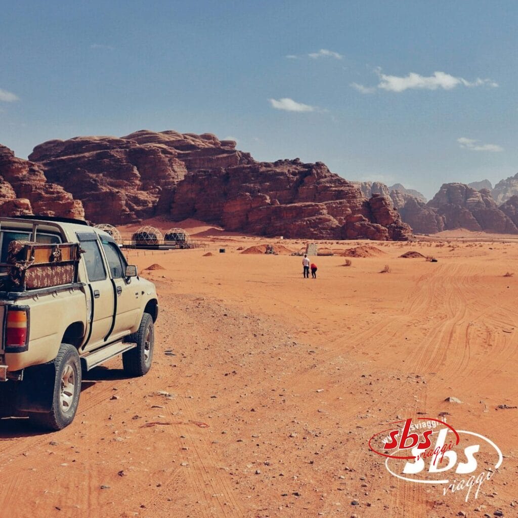 Un pick-up beige in un paesaggio desertico sabbioso con grandi formazioni rocciose, dove lo spirito della Tradizione incontra la Modernità del viaggio, e due persone camminano in lontananza.