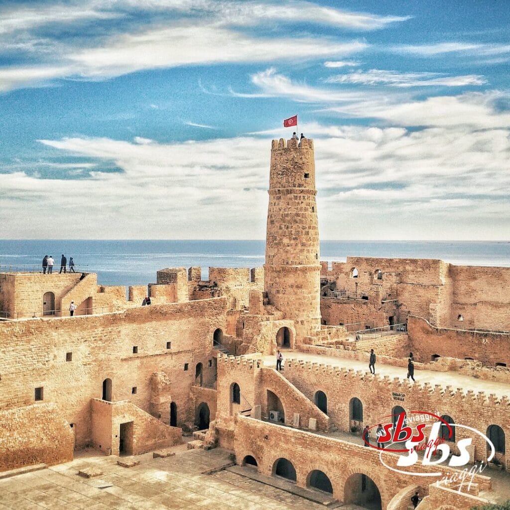Fortezza storica con una torre centrale su cui sventola una bandiera, che domina il mare sotto un cielo azzurro e nuvole: una tappa iconica di ogni avventura del Tour Autentica Tunisia.