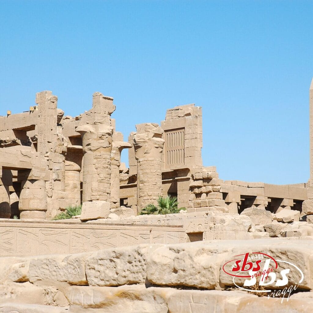 Antiche rovine in pietra con colonne sotto un cielo azzurro e terso evocano la grandiosità della Valle dei Re; logo