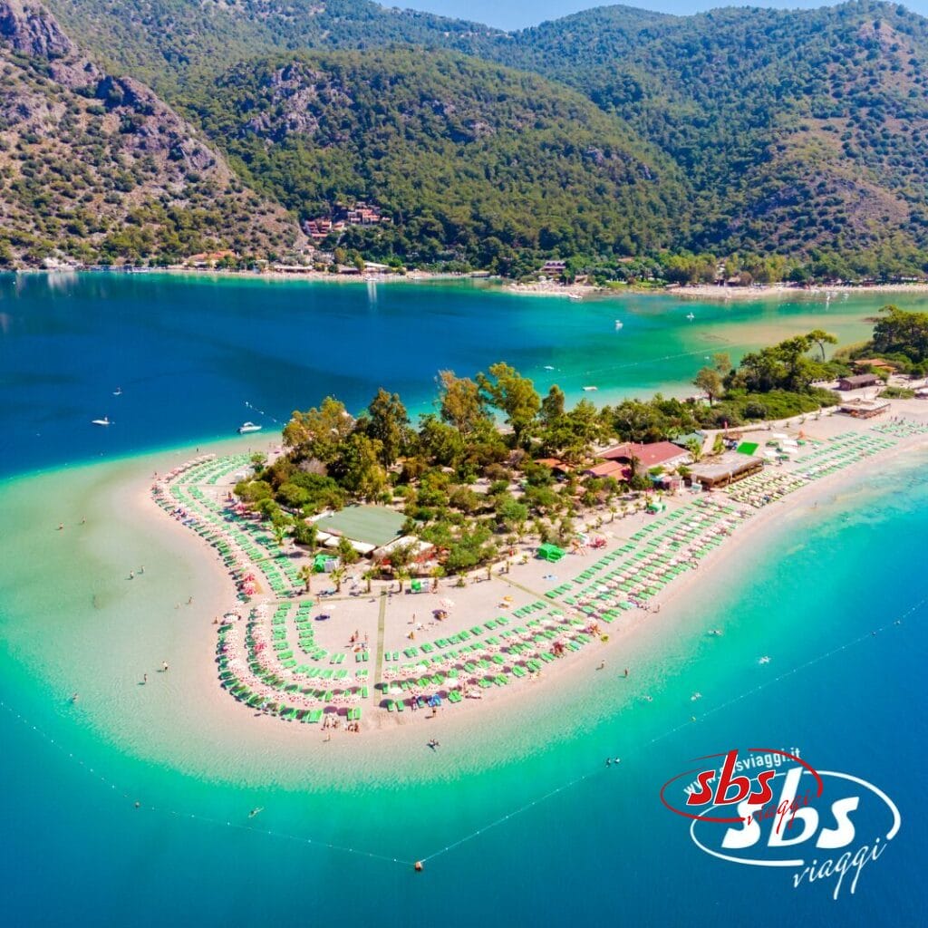 Vista aerea di una spiaggia sabbiosa con file di ombrelloni e lettini, circondata da acque turchesi e verdi colline, che evoca il fascino del Mediterraneo. Il logo 