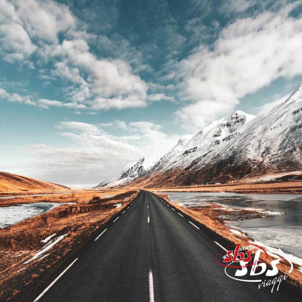 Strada dritta che conduce a montagne innevate sotto un cielo parzialmente nuvoloso, con campi e acqua su entrambi i lati, catturando la serena bellezza dell'autunno in Islanda, dove le cime lontane annunciano l'arrivo dell'inverno.