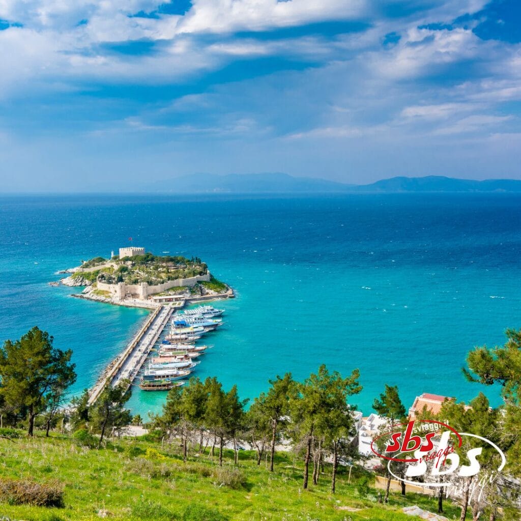 Vista di una piccola isola collegata alla terraferma da una strada rialzata, con un castello, barche ormeggiate, acqua turchese e alberi in primo piano sotto un cielo parzialmente nuvoloso: un luogo ideale per un Tour Kervansaray o un indimenticabile tour con mare.