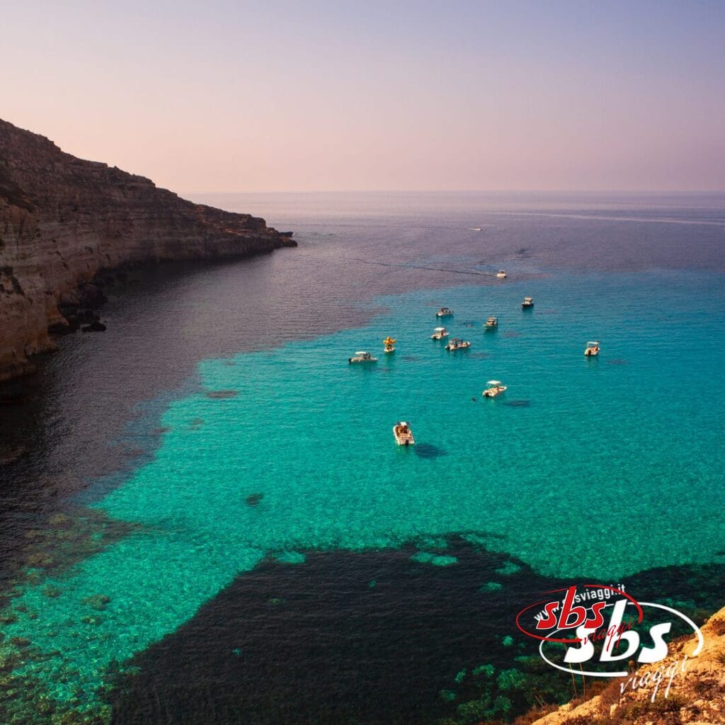 Piccole imbarcazioni galleggiano sulle limpide acque turchesi vicino alle scogliere rocciose, sotto un cielo velato, con il logo "sbs viaggi" e Case Vacanza Lampedusa ben visibili.