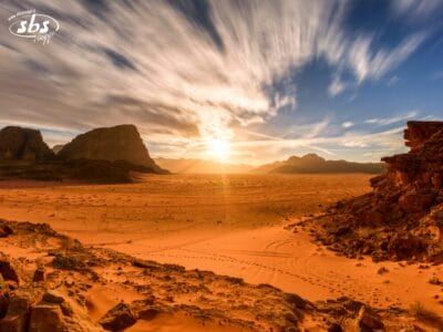 Un paesaggio desertico illuminato dal sole in Giordania, con sabbia arancione, formazioni rocciose e strisce di nuvole nel cielo: perfetto per un weekend indimenticabile alla scoperta di Petra.